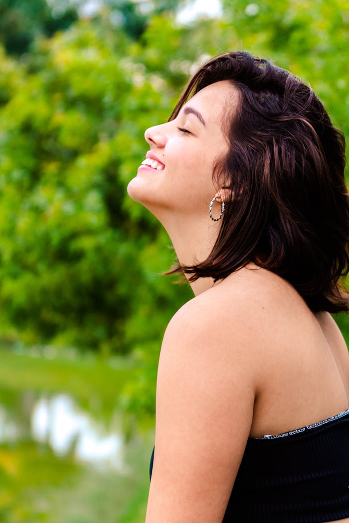 A joyful woman with short brunette hair smiles outdoors in a park, surrounded by lush greenery.